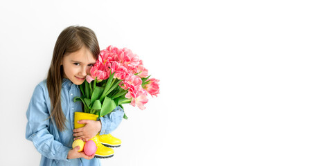 A long-haired lovely girl holds multicolored painted Easter eggs and a bouquet of pink tulips in her hands. Spring flowers stand in gumboots instead of a vase. 