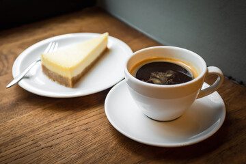 A cup of black coffee and a piece of cheesecake served on the plate, on the wooden windowsill, natural light