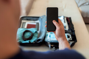 Young man packing clothes in suitcase, holding passport and immunization certificate. Person using...