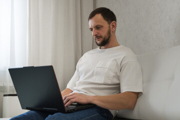 The concept of remote freelance work. A focused man with a beard looks at the laptop screen while sitting at home on the couch.