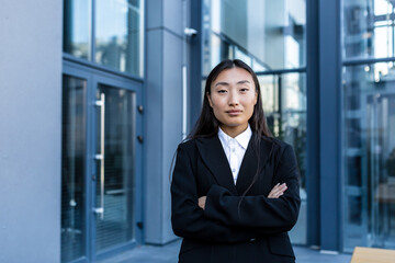 Successful business woman pensive and serious with crossed arms, outside the office, Asian woman looking at camera