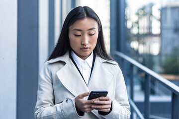 Beautiful Asian woman in business clothes uses the phone, walks near the modern office center outside