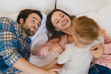 Parenthood concept. Caucasian young family of three parents mother and father with small little kid child toddler relaxing resting on the bed, playing and spending time together, taking care of kid