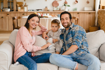 Happy caucasian cheerful young family of three looking at camera spending time together at home. Parents and toddler at home on the couch. Parenthood concept