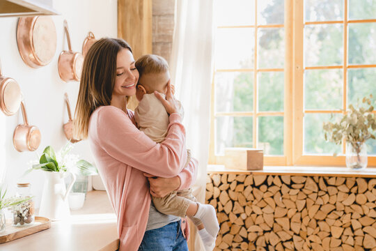 Happy Young Loving Mother Holding Hugging Embracing Little Small Kid Child Son Toddler, Nurturing, Singing Lullaby At Home Kitchen. Motherhood Concept