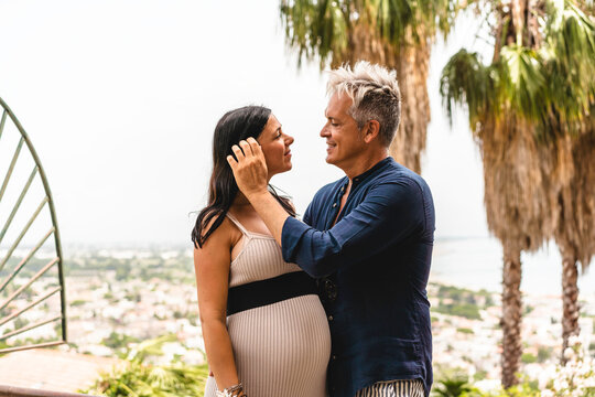 Couple, with pregnant woman, looking into each other's eyes and posing for a photo during summer vacation in the esotic garden - Cute wife and trendy middle aged man expecting a baby - Pregnancy life