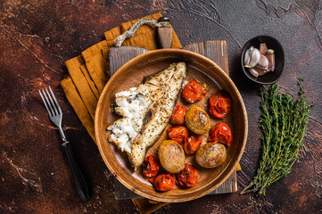 Baked halibut fish with roasted tomato and potato in wooden plate. Dark background. Top view