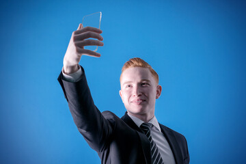 Portrait of a red-haired man in a suit with a smartphone in his hands on a blue background, taking a selfie. Business manager. Seller. Businessman. A successful businessman. Secretary. Assistant