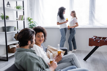 Fototapeta premium multiethnic lesbian couple holding bottles of beer and standing near window near blurred friends on foreground.