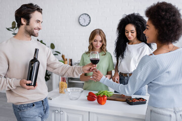 cheerful man giving glass of wine to african american woman near friends in kitchen.