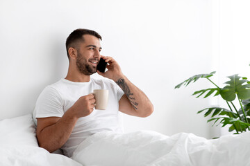 Smiling millennial muscular european male drinks coffee, calls by smartphone and enjoys good morning in bedroom