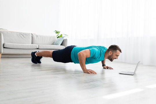 Cheerful millennial muscular european man doing push ups and watching online lesson on computer in living room