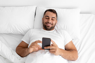 Cheerful millennial muscular european man typing on phone, chatting, searching and surfing in internet, lies on bed