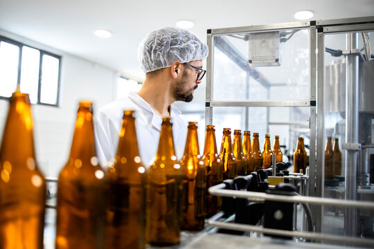 Factory worker working in alcohol beverage bottling factory. Technologist standing by production line machine and controlling beer bottles.