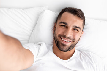 Smiling glad millennial muscular caucasian man waking up taking selfie on white bed on soft pillow...