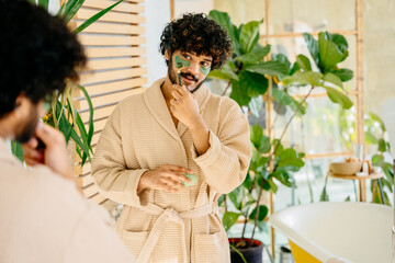 Charismatic hispanic man in beige bathrobe enjoy morning routine in cozy bathroom interior with mirror and green plants. Cosmetology beauty and lifestyle concept - obrazy, fototapety, plakaty
