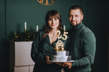Stylish bearded man and beautiful brunette girl hold a delicious birthday cake with numbers 25 in...