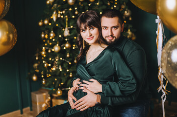A stylish, smiling bearded man and a beautiful pregnant brunette girl sit and gently hug against the background of a decorated Christmas tree, balloons.
