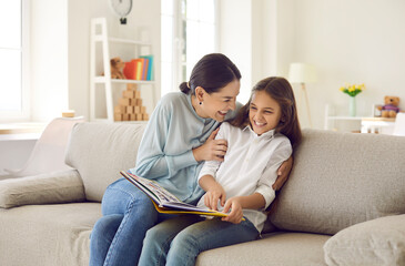 Smiling young Caucasian mother and teen daughter sit relax on sofa laugh reading book together....