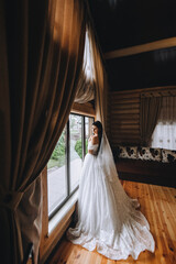 A beautiful bride in a long white dress stands in a wooden house, interior near a large window. Wedding photography, portrait.