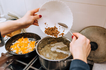 A woman, a cook, prepares soup from vegetables in boiling water, pouring buckwheat from a plate into a saucepan. Cooking food in the kitchen.