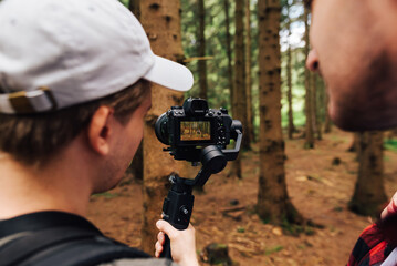 Young man holds a camera on the stabilizer and shows a colleague the video result on the camera screen. Creating a travel video. Video travel content