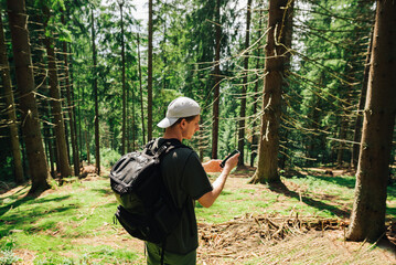 Fototapeta premium Young tourist in casual clothes and with a backpack on his back uses the Internet on a smartphone in the middle of a mountain coniferous forest.