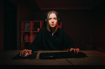 Attractive girl gamer sitting at home at the computer in a headphone and playing online games, looking intently at the camera. © bodnarphoto
