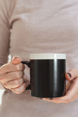 Black coffee mug mockup for design demonstration. Stylish cup mock up in female hands on white wall background.