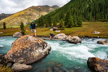 Young man and woman crossing wild turquoise river on tiny wooden bridge  on trail to Ala Kul lake....