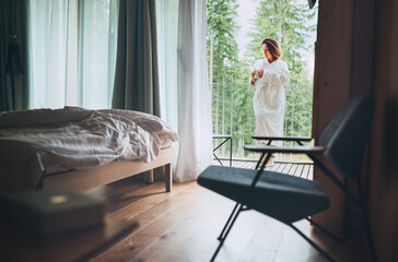 Woman dressed white bathrobe standing on forest house balcony and enjoying fresh air and morning teacup. Inside a Scandinavian interior design room view with a not made bed. Cozy living concept image. - obrazy, fototapety, plakaty