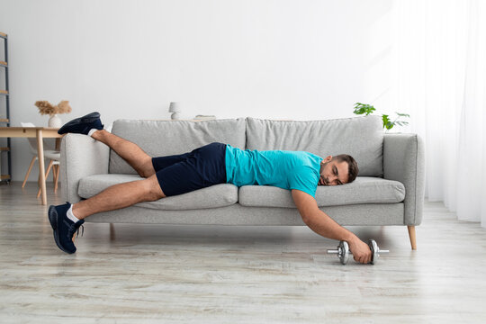 Tired funny millennial muscular european man lies on sofa with dumbbell after training in living room