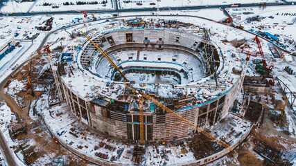 Round stadium construction site covered with snow with high cranes and machine equipment at building finishing process aerial view