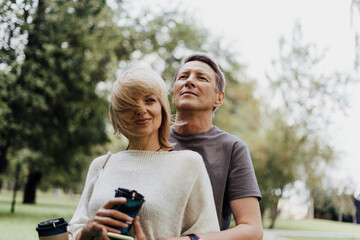 An adult mature happy couple in love hugging outdoors in city park. A blonde caucasian man and woman spend time together and drinking coffee. Senior wife and husband walking outside