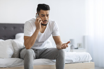 Portrait Of Worried Young Arab Man Talking On Cellphone In Bedroom