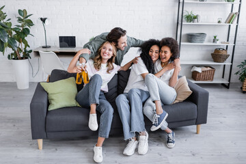 happy man holding bottles of beer and hugging interracial women on sofa.