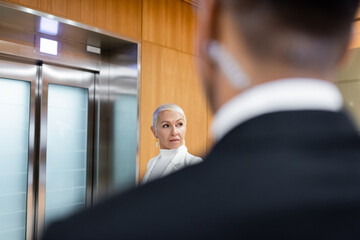 senior business lady waiting for elevator in hotel near african american bodyguard on blurred...