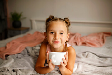 Cute little happy girl with two ponytails in her bed drinking hot tea from white cup