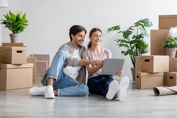 Fototapeta na wymiar Smiling caucasian young husband and wife look at pc among cardboard boxes with stuff in room