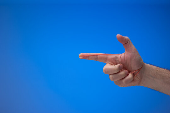 Male hand making a pistol gesture with am elastic rubber band. Close up studio shot, isolated on blue background