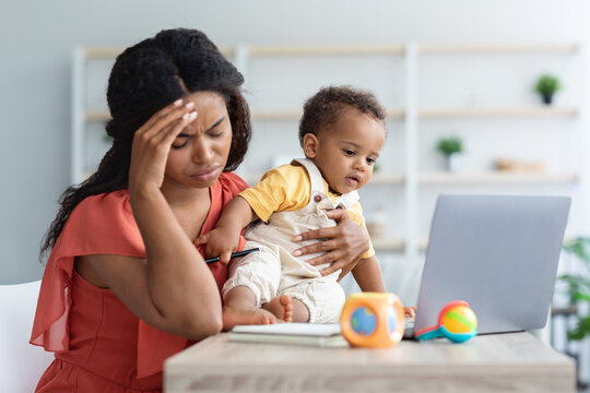 Freelance Stress. Exhausted Black Woman Working On Laptop With Baby In Arms