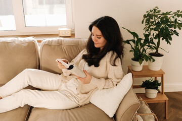 A pretty young female student is reading a book studying while resting on a sofa in a cozy Scandinavian-style living room in an apartment. Domestic life. Selective focus