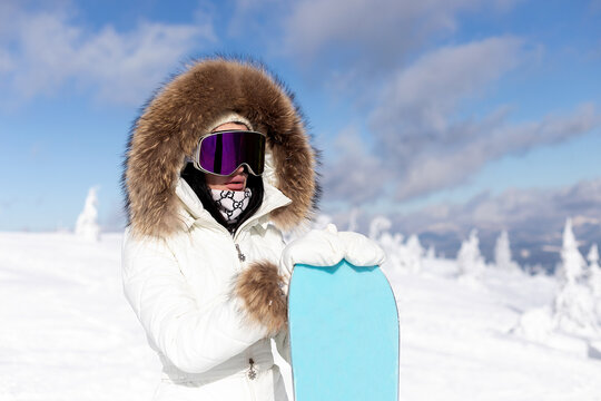 winter, leisure, sport and people concept - Girl snowboarder stands with snowboard on mountain's top on sunrise backdrop. woman in fur hood and snowboarding goggles