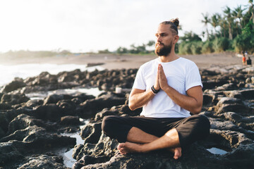 Naklejka na ściany i meble Harmony and meditation training during morning yoga at coastline with rocks, Caucaisan male in sportswear sitting in lotus pose during hatha practice for exercising body concentration in asana