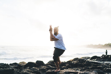 Flexible man practice balance asana breathing in pranayama at morning coastline, concentrated man standing at one leg on stone doing stretching exercises during workout for feeling vitality