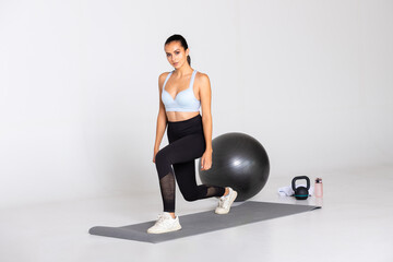 female workout with lunges and hands on her side on a grey mat with gym equipment on white background in sports wear, trainer looks into the camera during workout and stretch on mat
