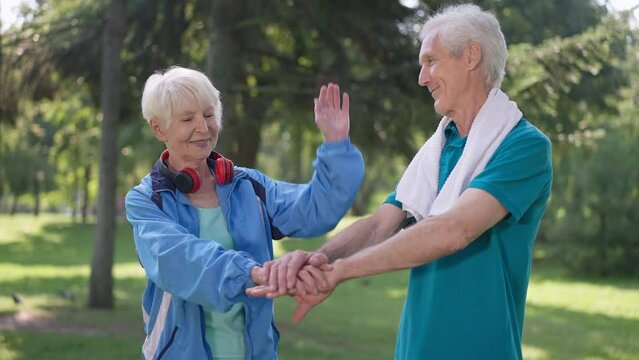 Medium shot of positive senior couple stacking hands in slow motion smiling standing in park outdoors. Happy fit confident Caucasian husband and wife enjoying training on sunny day