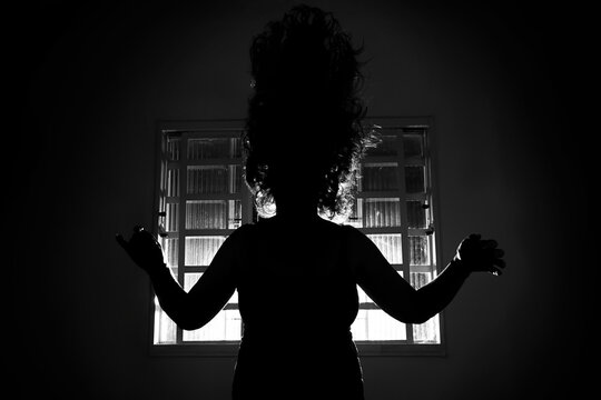 Silhueta sinistra de uma pessoa em contraluz na frente de uma janela em um quarto escuro.