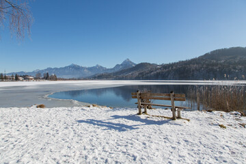 Idyllic sunny winter landscape at lake Weißensee with an inviting wooden bench to enjoy the beautiful panorama view to the Füssen mountains and the twin peak mountain Säuling near the Austrian border