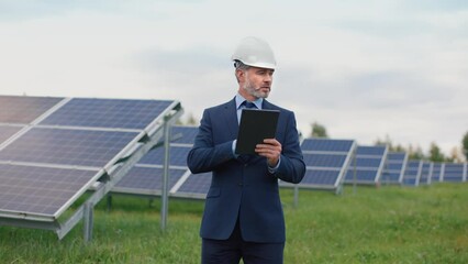 Successful Caucasian male businessman in suit and hard hat stands among field with solar panels inspecting and checking data in tablet. - Powered by Adobe